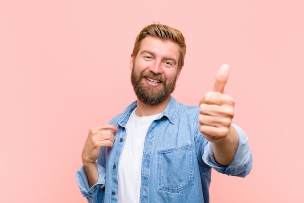 Giovane uomo adulto biondo sentirsi orgoglioso, spensierato, sicuro e felice, sorridendo positivamente con i pollici in su