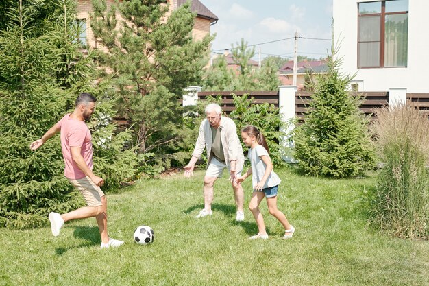 Giovane uomo adulto che gioca a calcio con il padre anziano e la figlia piccola sul prato in cortile