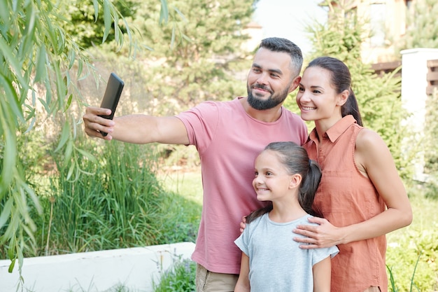 Giovane uomo adulto che tiene in mano uno smartphone che scatta selfie di lui, sua moglie e sua figlia nel cortile sul retro in una soleggiata giornata estiva