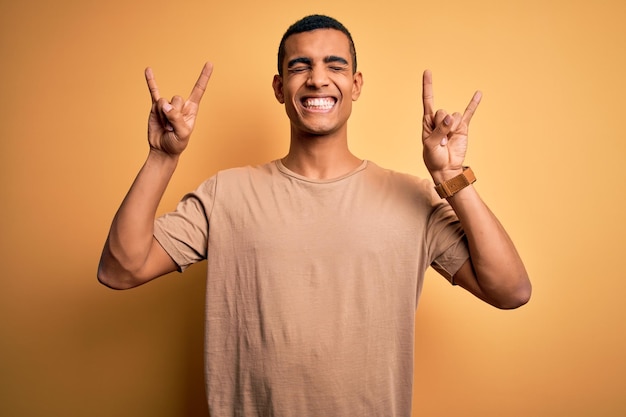 Giovane uomo affascinante afroamericano che indossa una maglietta casual in piedi su uno sfondo giallo urlando con un'espressione pazza facendo il simbolo del rock con le mani in alto stella musicale concetto di musica pesante