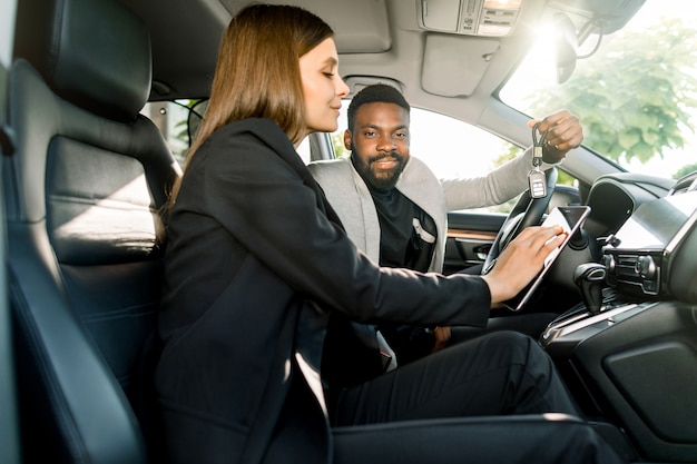 Giovane uomo africano allegro bello che tiene le chiavi dell'automobile dalla sua nuova automobile di lusso, mentre sedendosi nell'automobile insieme al rappresentante di automobile grazioso della donna che tiene una compressa