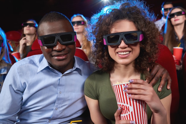 Giovane uomo africano felice che abbraccia la sua bella fidanzata allegra mentre si gode la visione di un film in 3D al cinema mangiando insieme relazioni di popcorn che frequentano la tecnologia del tempo libero della gente moderna.