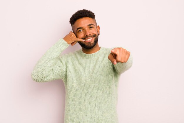 Giovane uomo afro che sorride allegramente e indica la fotocamera mentre si effettua una chiamata in seguito gesto parlando al telefono