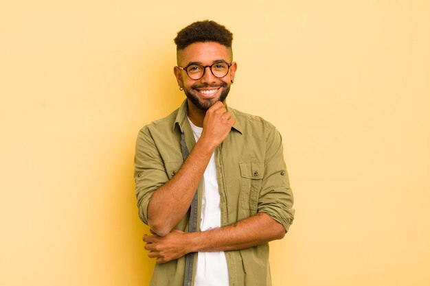 Giovane uomo afro sorridente godendosi la vita sentendosi felice, amichevole, soddisfatto e spensierato con la mano sul mento