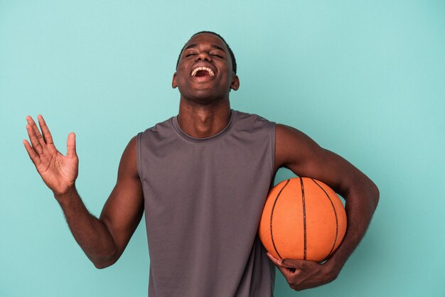 Giovane uomo afroamericano che gioca a basket isolato su sfondo blu ricevendo una piacevole sorpresa, eccitato e alzando le mani.