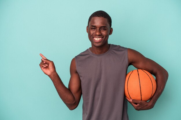 Giovane uomo afroamericano che gioca a basket isolato su sfondo blu sorridente e indicando da parte mostrando qualcosa in uno spazio vuoto