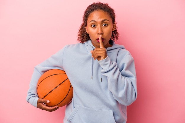 Giovane uomo afroamericano che gioca a basket isolato su sfondo rosa mantenendo un segreto o chiedendo silenzio.