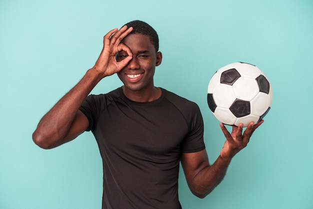 Giovane uomo afroamericano che gioca a calcio isolato su sfondo blu eccitato mantenendo il gesto giusto sull'occhio