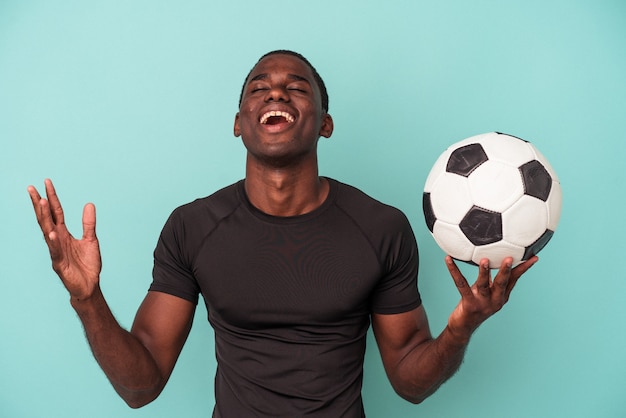 Giovane uomo afroamericano che gioca a calcio isolato su sfondo blu riceve una piacevole sorpresa, eccitato e alzando le mani.