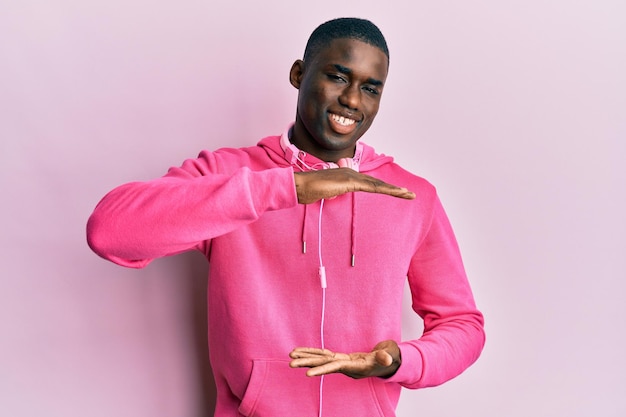 Giovane uomo afroamericano che indossa abiti da palestra e utilizza le cuffie gesticolando con le mani che mostrano il simbolo di misura del segno di grandi e grandi dimensioni sorridendo guardando il concetto di misurazione della fotocamera