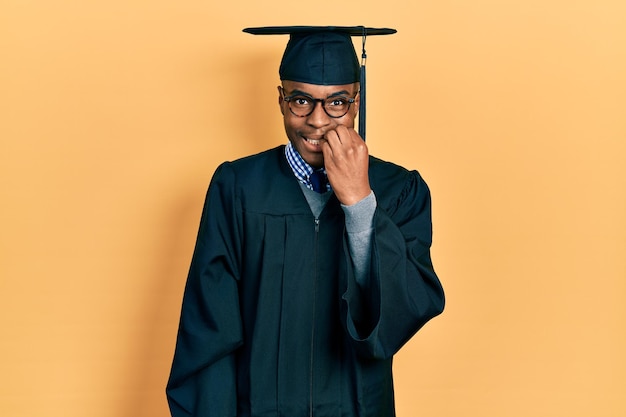 Giovane uomo afroamericano che indossa il berretto di laurea e l'abito da cerimonia che sembra stressato e nervoso con le mani sulla bocca che mordono il problema dell'ansia delle unghie