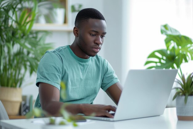 Giovane uomo afroamericano che indossa jeans e una maglietta verde seduto a lavorare al portatile in un elegante ufficio domestico bianco