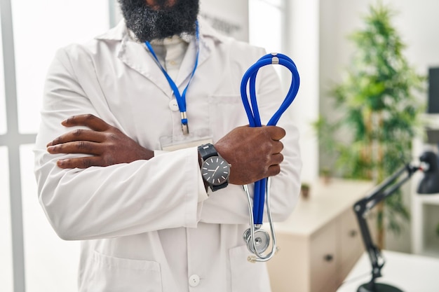 Giovane uomo afroamericano che indossa l'uniforme del medico in piedi con le braccia incrociate gesto alla clinica