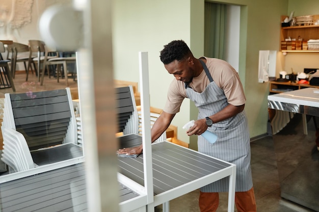 Giovane uomo afroamericano che indossa un grembiule che inizia la giornata lavorativa nella moderna caffetteria preparando i tavoli per i clienti che li puliscono prima dell'apertura