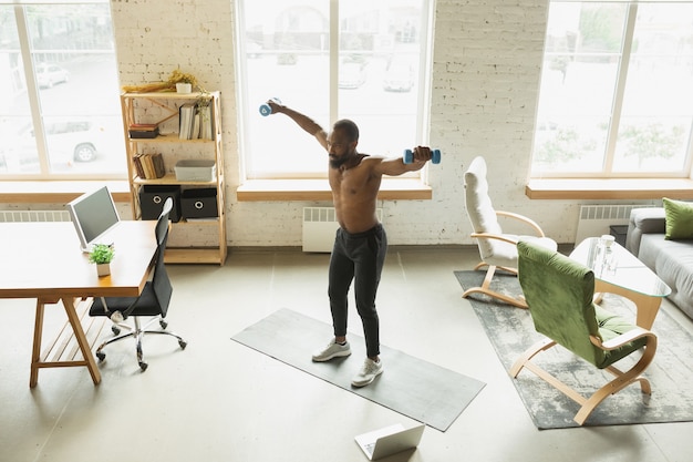 Giovane uomo afroamericano che si allena a casa durante la quarantena dell'epidemia di coronavirus, esercizi di fitness, aerobica. Rimanere sportivo durante l'isolamento. Benessere, concetto di movimento. Pesi in su.