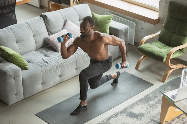 Giovane uomo afroamericano che si allena a casa durante la quarantena dell'epidemia di coronavirus, esercizi di fitness, aerobica. Rimanere sportivo durante l'isolamento. Benessere, concetto di movimento. Siediti.