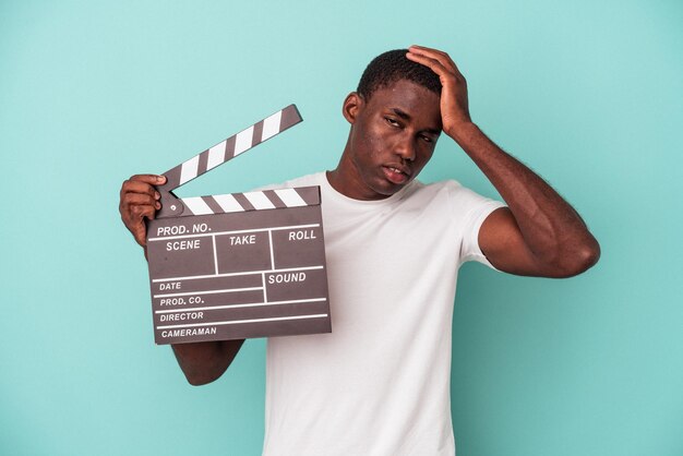 Giovane uomo afroamericano che tiene clapperboard isolato su sfondo blu essendo scioccato, ha ricordato un incontro importante.