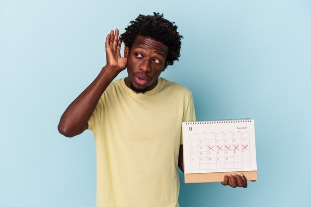 Giovane uomo afroamericano che tiene il calendario isolato su sfondo blu cercando di ascoltare un pettegolezzo.