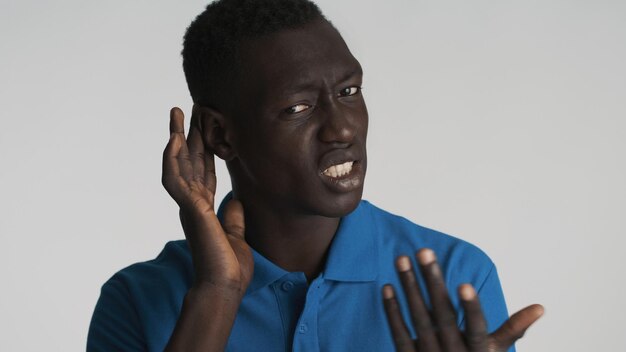 Giovane uomo afroamericano che tiene la mano vicino all'orecchio chiedendo di nuovo sulla fotocamera su sfondo bianco. Non sentire il gesto