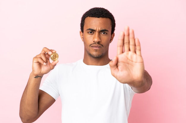 Giovane uomo afroamericano che tiene un Bitcoin su sfondo rosa isolato che fa un gesto di arresto