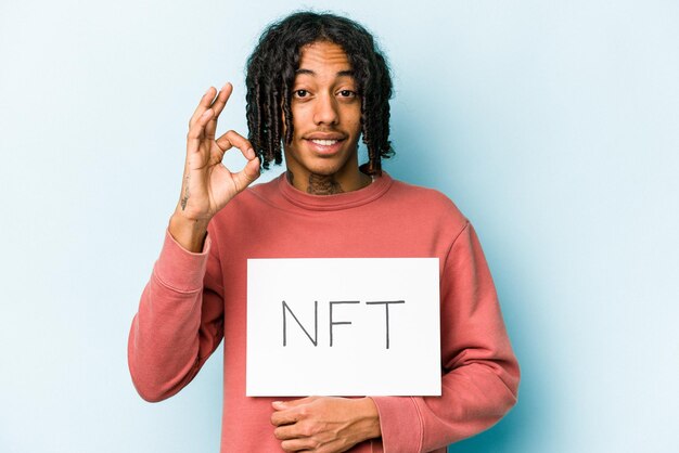 Giovane uomo afroamericano con cartello NFT isolato su sfondo blu allegro e fiducioso che mostra un gesto ok