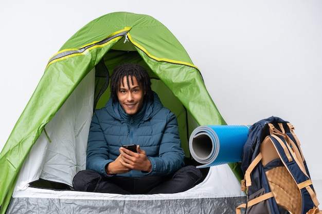 Giovane uomo afroamericano dentro una tenda verde di campeggio che invia un messaggio con il cellulare