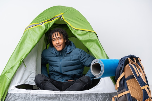 Giovane uomo afroamericano dentro una tenda verde di campeggio che posa con le armi all'anca e a sorridere