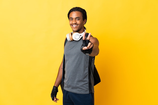 Giovane uomo afroamericano di sport con le trecce con la borsa isolata su fondo giallo che sorride e che mostra il segno di vittoria