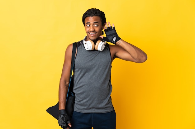 Giovane uomo afroamericano di sport con le trecce con la borsa sul giallo che ascolta qualcosa mettendo la mano sull'orecchio