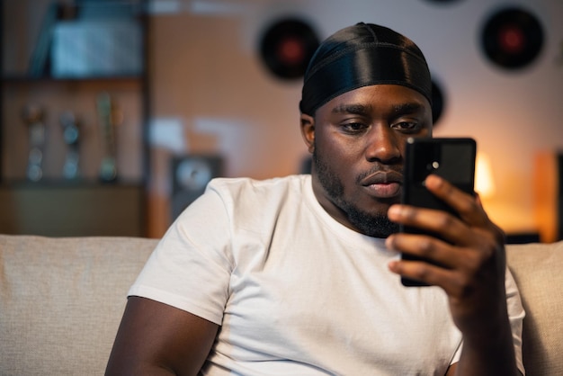 Giovane uomo afroamericano in possesso di smartphone che invia messaggi di testo o gioca a giochi mobili seduto sul divano