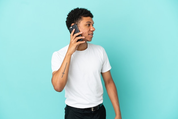 Giovane uomo afroamericano isolato su sfondo blu che tiene una conversazione con il telefono cellulare con qualcuno
