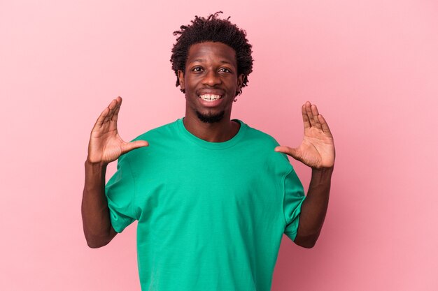 Giovane uomo afroamericano isolato su sfondo rosa che tiene qualcosa con le palme, offrendo alla macchina fotografica.