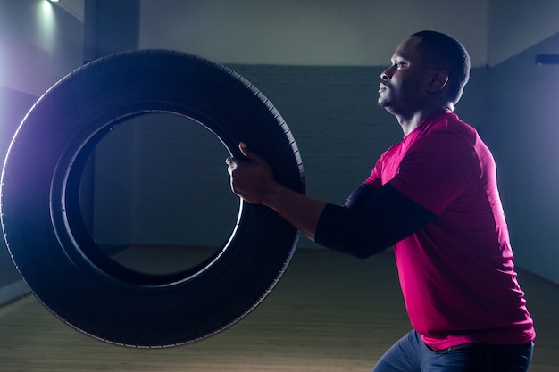 Giovane uomo afroamericano muscolare senza camicia in pneumatico sportivo che si esercita con pneumatico in palestra su uno sfondo nero nell'officina dello studio