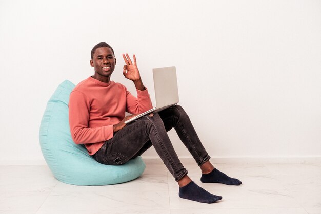 Giovane uomo afroamericano seduto su un puff utilizzando laptop isolato su sfondo bianco allegro e fiducioso che mostra gesto ok.