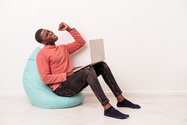 Giovane uomo afroamericano seduto su un puff utilizzando laptop isolato su sfondo bianco alzando il pugno dopo una vittoria, concetto vincitore.