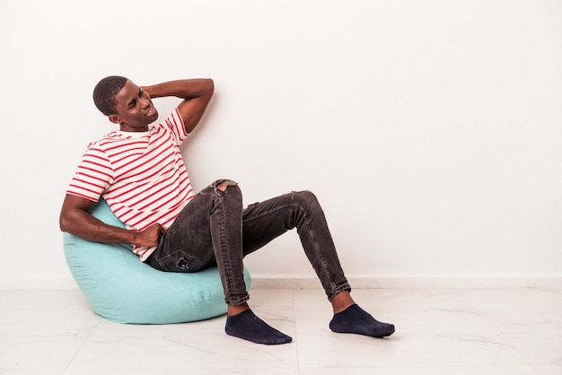 Giovane uomo afroamericano seduto su un soffio isolato su sfondo bianco toccando la parte posteriore della testa, pensando e facendo una scelta.