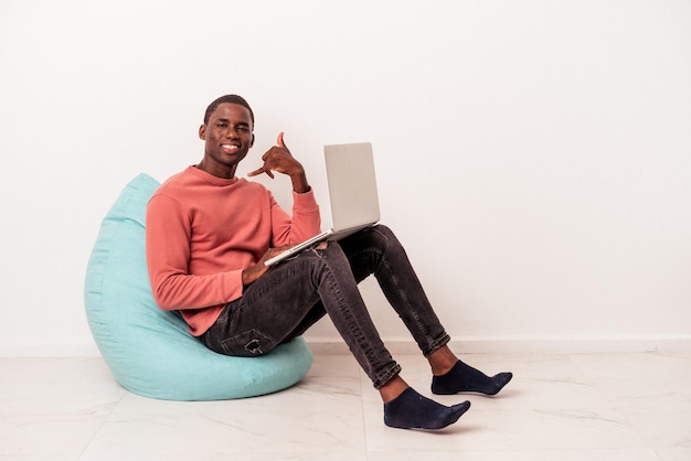 Giovane uomo afroamericano seduto su un soffio utilizzando un laptop isolato su sfondo bianco che mostra un gesto di chiamata di telefonia mobile con le dita.