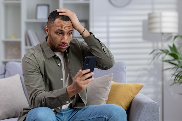 Giovane uomo afroamericano seduto sul divano di casa e guardando il telefono sotto shock tenendosi per mano