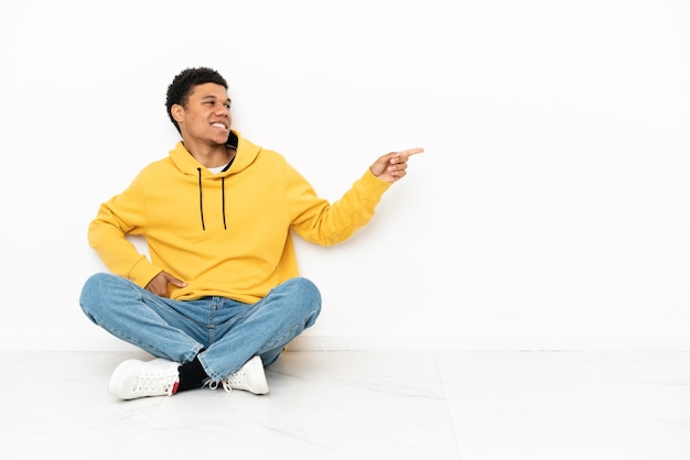 Giovane uomo afroamericano seduto sul pavimento isolato su sfondo bianco che punta il dito di lato e presenta un prodotto