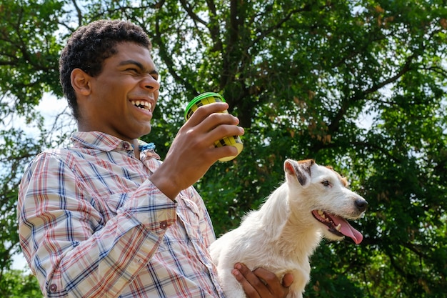 Giovane uomo afroamericano sorridente che tiene cane e caffè del terrier di Jack Russell per andare. Camminare nel parco in estate, vista dal basso