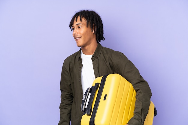 Giovane uomo afroamericano sulla parete porpora in vacanza con la valigia di viaggio