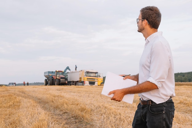 Giovane uomo agronomo in piedi sul campo di grano che controlla la qualità mentre la mietitrebbia lavora.