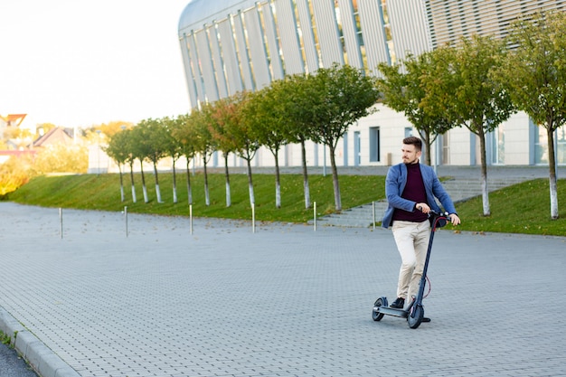 Giovane uomo alla moda bello con il motorino elettronico che sta sul marciapiede vicino all'aeroporto sul tramonto