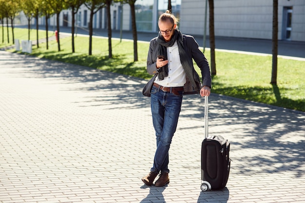 Giovane uomo alla moda che per mezzo del suo smartphone mentre stando con l'uscita vicina della valigia l'aeroporto all'aperto. Stile aziendale, viaggiatore, stile di vita moderno, viaggio d'affari.