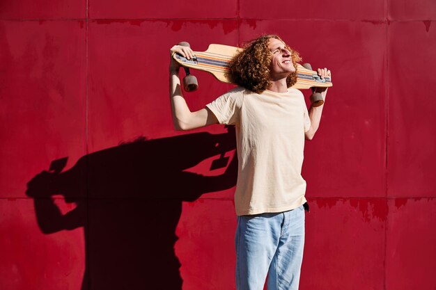 giovane uomo alla moda con i capelli ricci che guarda il sole con il suo skateboard sulle spalle e con uno sfondo rosso