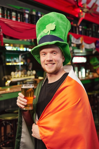 Giovane uomo allegro con un grande cappello verde che ti guarda con un sorriso mentre brinda con un bicchiere di birra davanti alla telecamera contro il bancone del bar