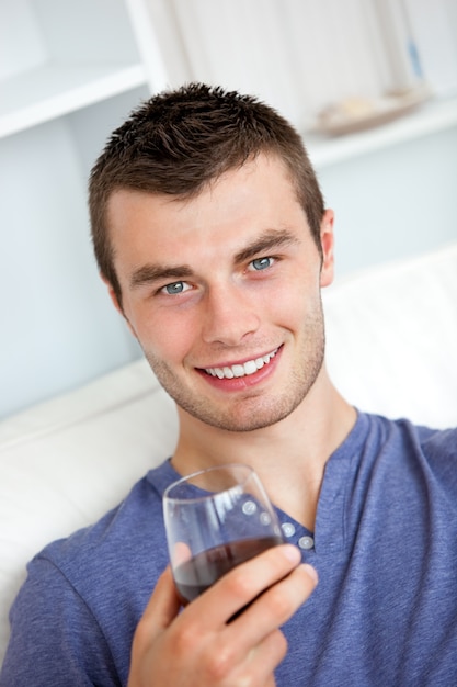 Giovane uomo animato bere vino guardando la telecamera seduta sul divano