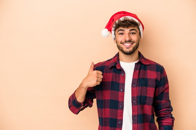 Giovane uomo arabo che celebra il Natale isolato su fondo beige che sorride e che alza pollice su