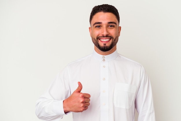 Giovane uomo arabo che indossa abiti arabi tipici isolati su sfondo bianco sorridendo e alzando il pollice thumb