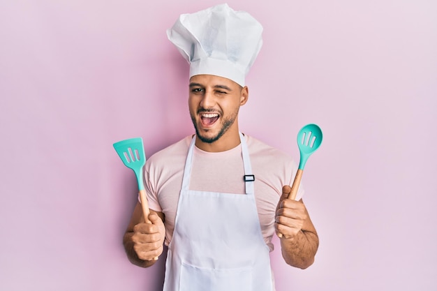 Giovane uomo arabo che indossa un grembiule da cuoco professionale e un cappello che tiene un cucchiaio che strizza l'occhio guardando la telecamera con un'espressione sexy faccia allegra e felice